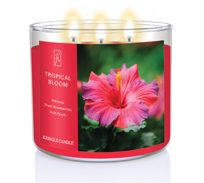 A three-wick lit candle in a clear glass jar with a vibrant floral-themed label. The candle’s wax is hot pink, and all three wicks are burning.