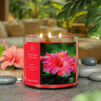 A three-wick lit candle in a clear glass jar with a vibrant floral-themed label. The candle’s wax is hot pink, and all three wicks are burning.