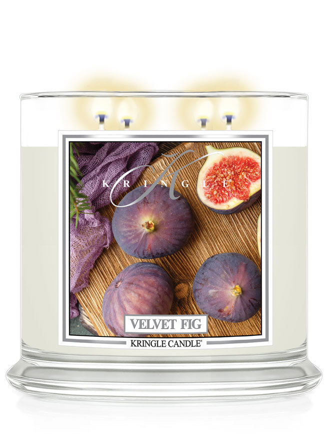 Lit 4-wick candle jar with label showing whole and halved figs on a wooden surface, next to a woven purple cloth.