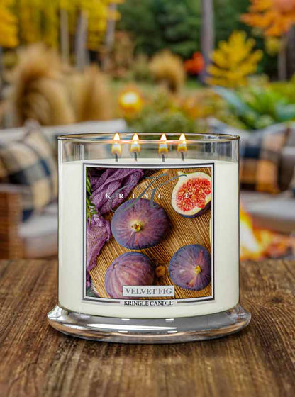 Lit 4-wick candle jar with label showing whole and halved figs on a wooden surface, next to a woven purple cloth.